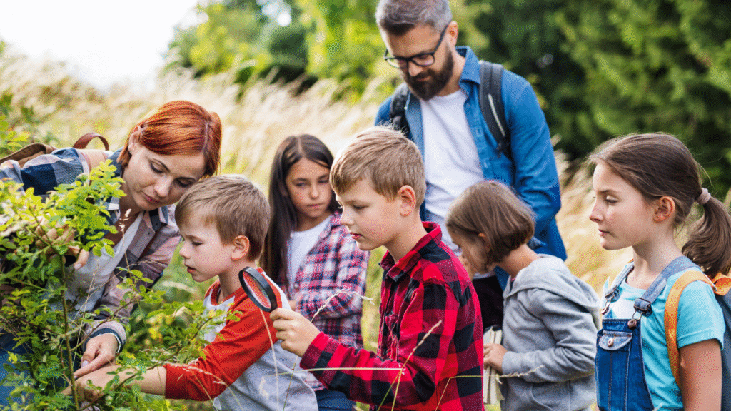 Teacher guiding curious elementary and middle school students during an outdoor nature activity, with kids exploring plants and using magnifying glasses during hands-on learning in a project-based school setting.