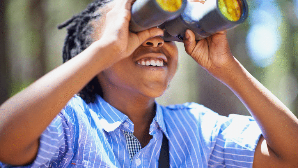 Smiling child exploring nature with binoculars during outdoor learning, developing curiosity and observational skills in a sensory-friendly, hands-on education setting.