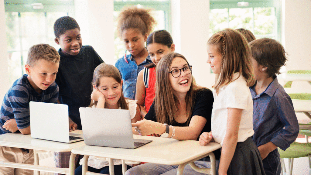 Smiling teacher supporting diverse elementary students working on laptops in a bright, inclusive classroom at a flexible, neurodiversity-affirming school